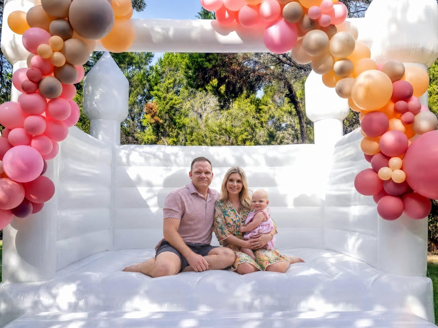 A family of three sitting together inside a white Hartwell Inflatables castle bounce house beneath a blush and peach balloon arch